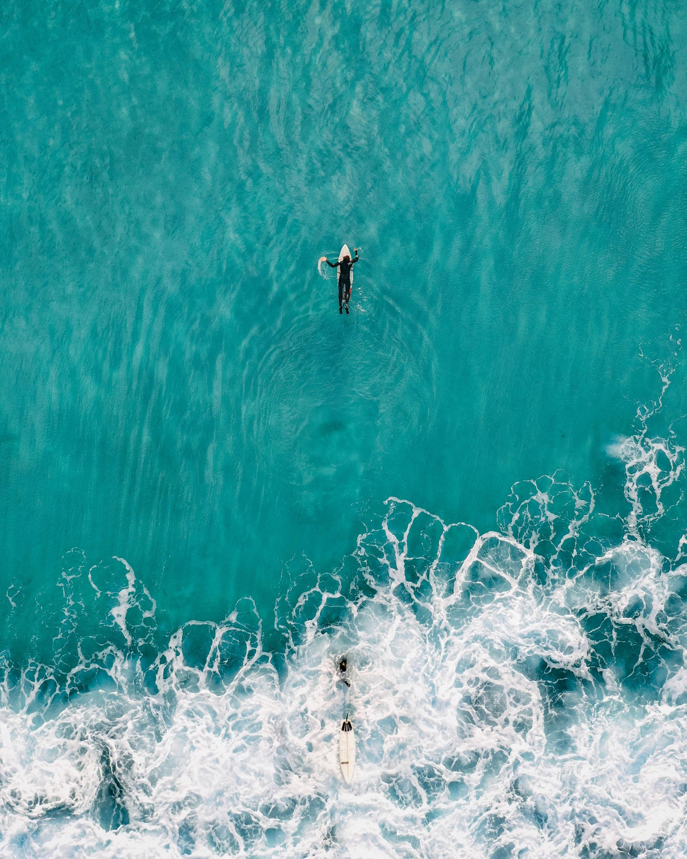 A surfer paddles out over clear turquoise water, seen from above
